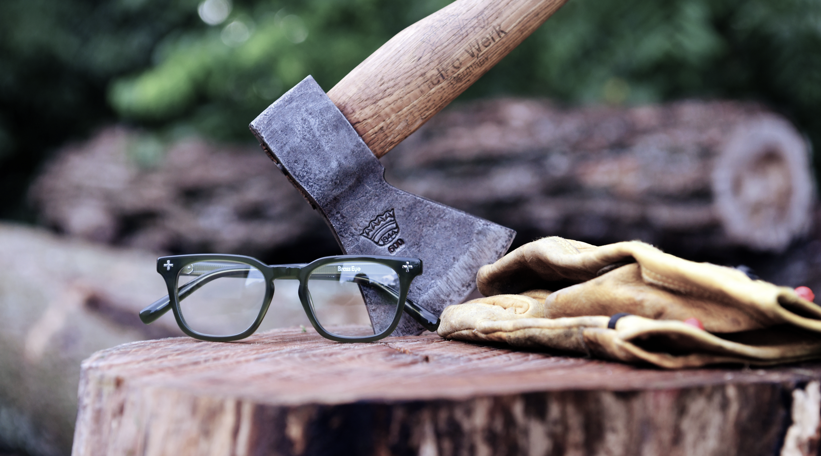 A pair of moss Brass Eye quality safety glasses resting on a wooden stump next to a leather work glove and a hatchet with a wooden handle, with logs and blurred greenery in the background.