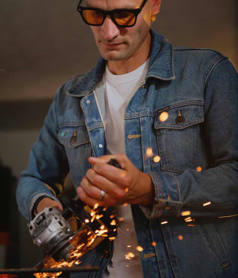 A man wearing Brass Eye quality safety glasses with oil colored lenses, a white t-shirt, and a blue denim jacket operates a grinder, creating bright sparks in a workshop setting.