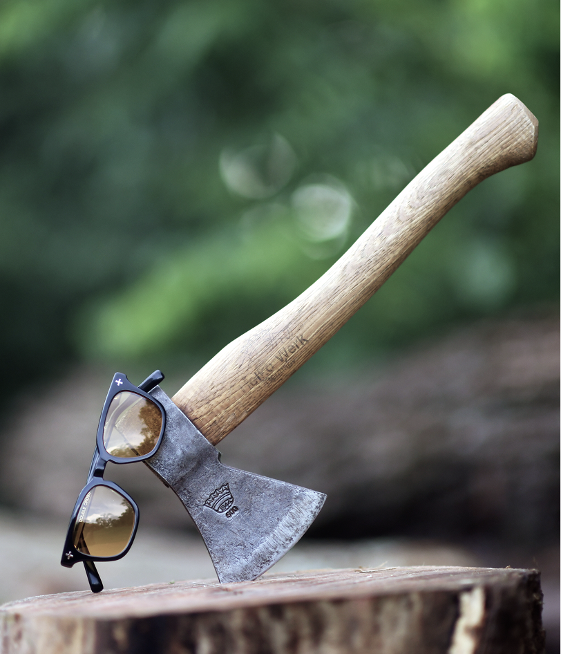 A small hatchet with a light wood handle and a weathered metal blade is embedded in a tree stump outdoors. A pair of Brass Eye quality safety glasses rests on the blade.