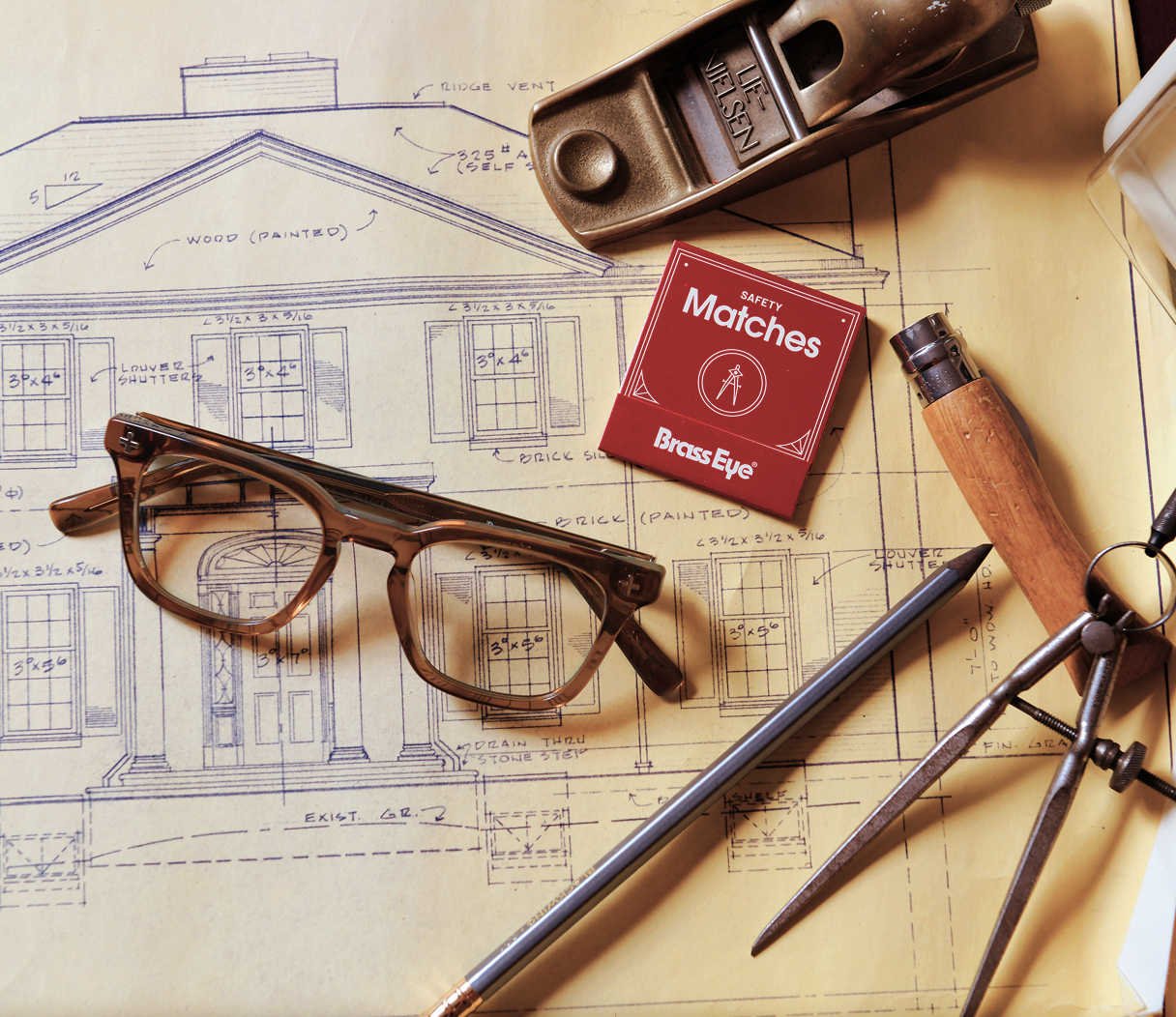A styled flat-lay of vintage architectural tools and Brass Eye quality safety glasses on top of house sketch blueprints. The set includes brown acetate eyeglasses, a red box of Brass Eye matches, a wooden-and-metal carpenter's square, a metal plane, and a pencil on a yellow paper background.
