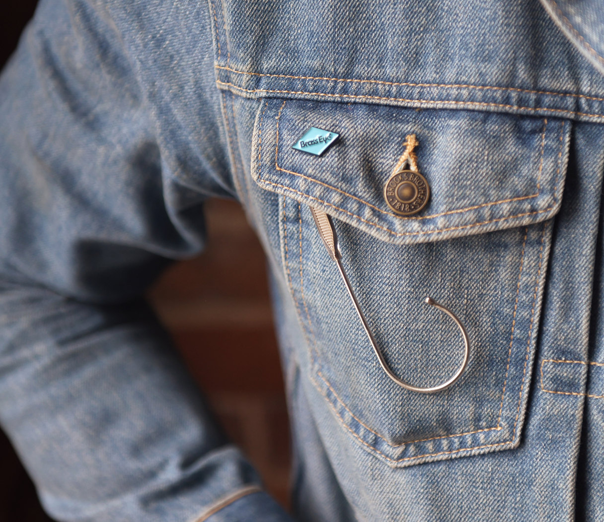 Close-up of a blue denim jacket with a chest pocket, featuring a small blue diamond-shaped Brass Eye branded pin, and a silver fishhook accessory.
