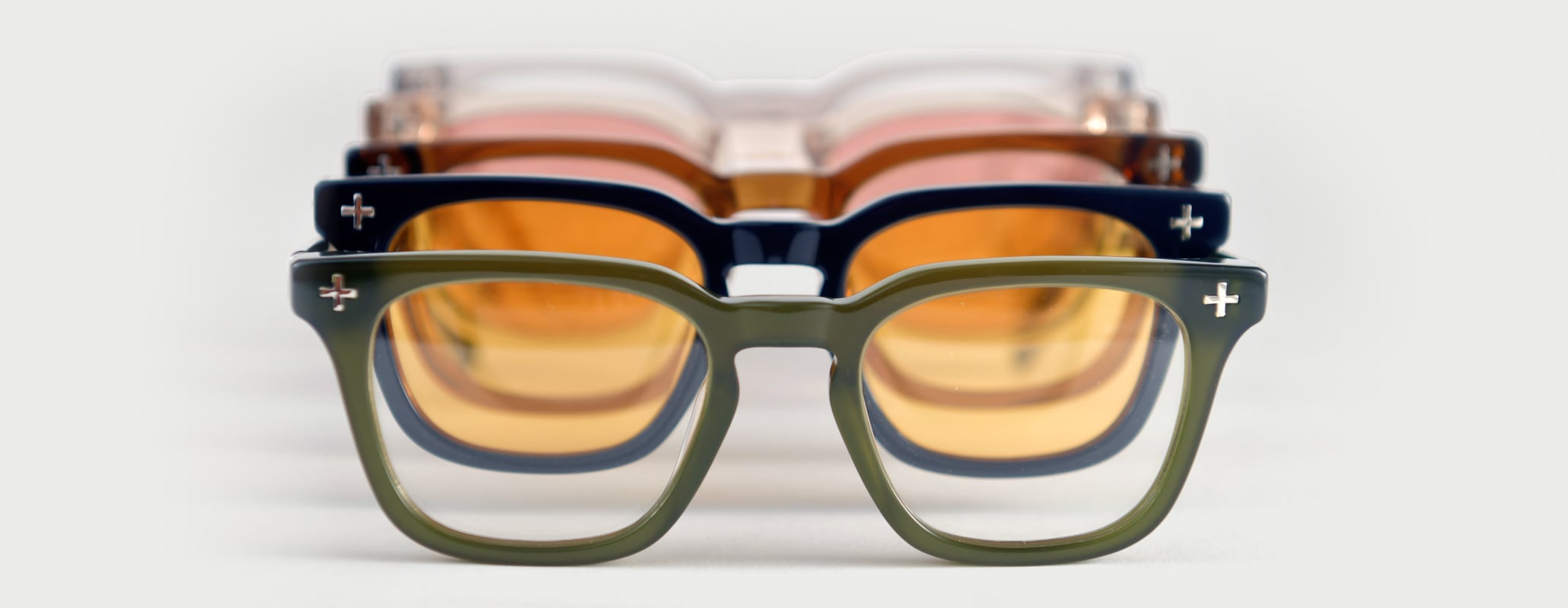 Three pairs of Brass Eye quality safety glasses stacked together on a light gray background. The front pair has an olive green frame with amber-tinted lenses, the middle pair has a dark navy blue frame with amber-tinted lenses, and the back pair has a translucent pink frame with amber-tinted lenses.