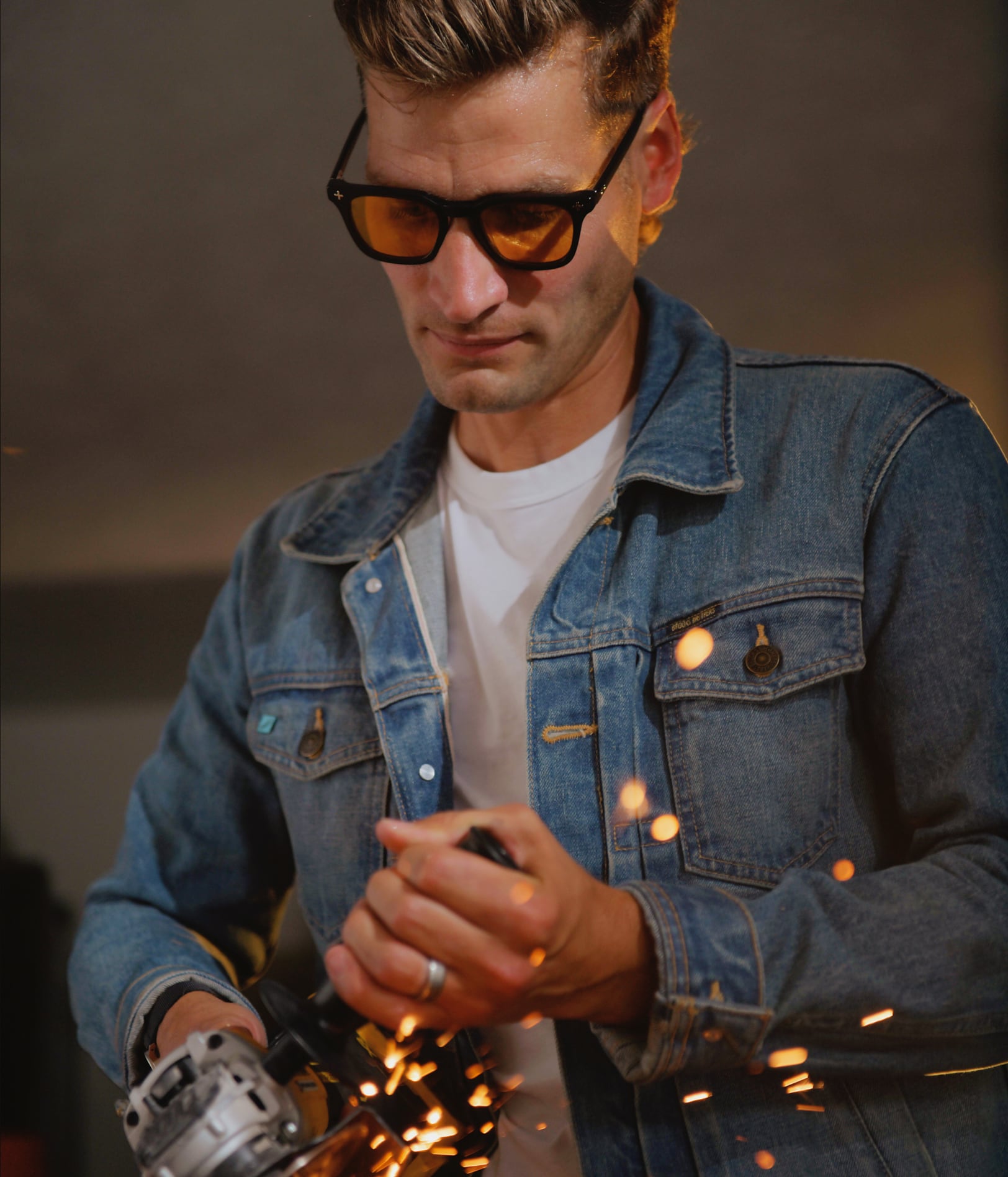 A man wearing Brass Eye quality safety glasses with amber lenses and a blue denim jacket works with sparks flying, holding a metal angle grinder.