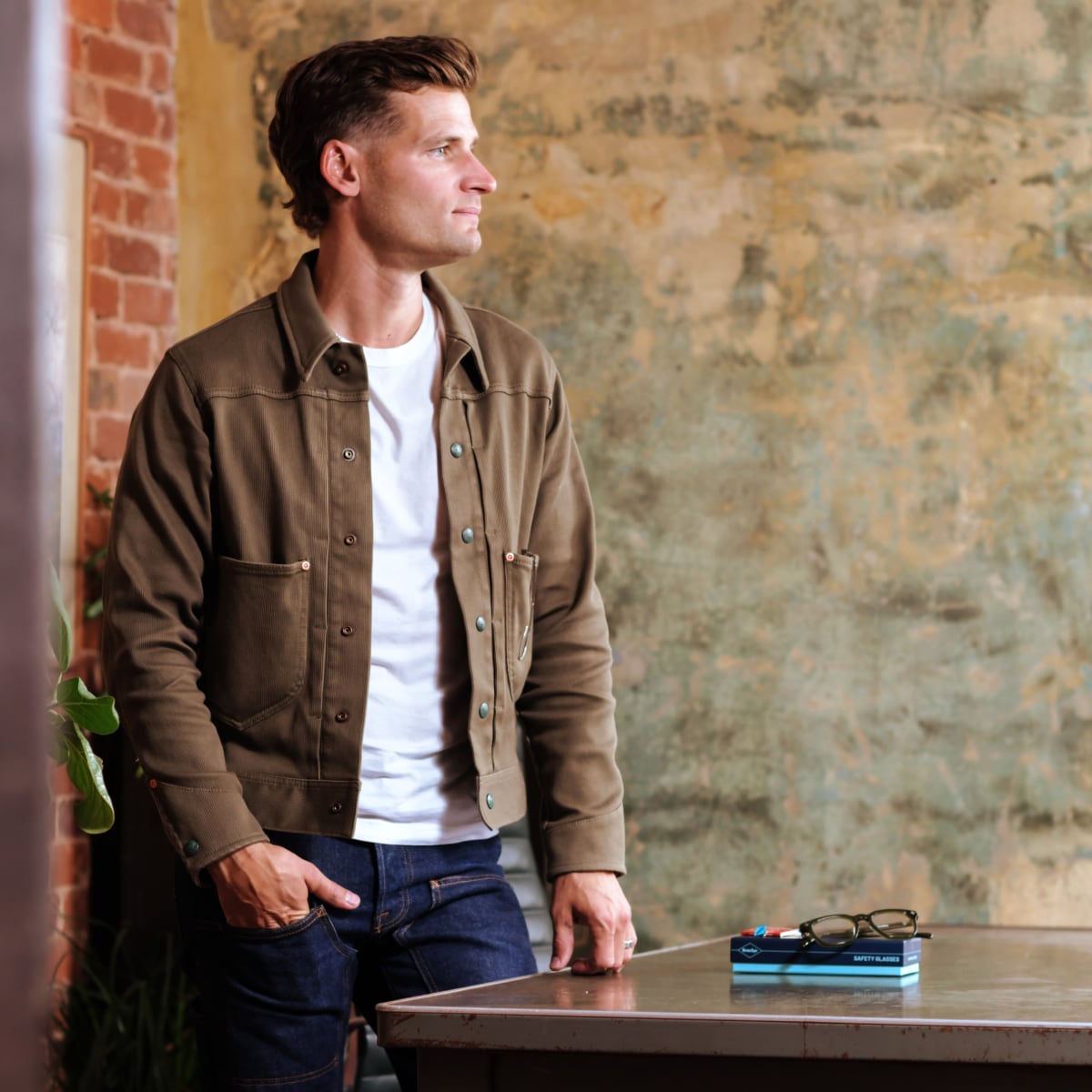 A man wearing a brown utility-style overshirt over a white t-shirt and blue jeans, standing in a rustic room with a brick column and textured wall next to a table with Brass Eye quality safety glasses and a book.