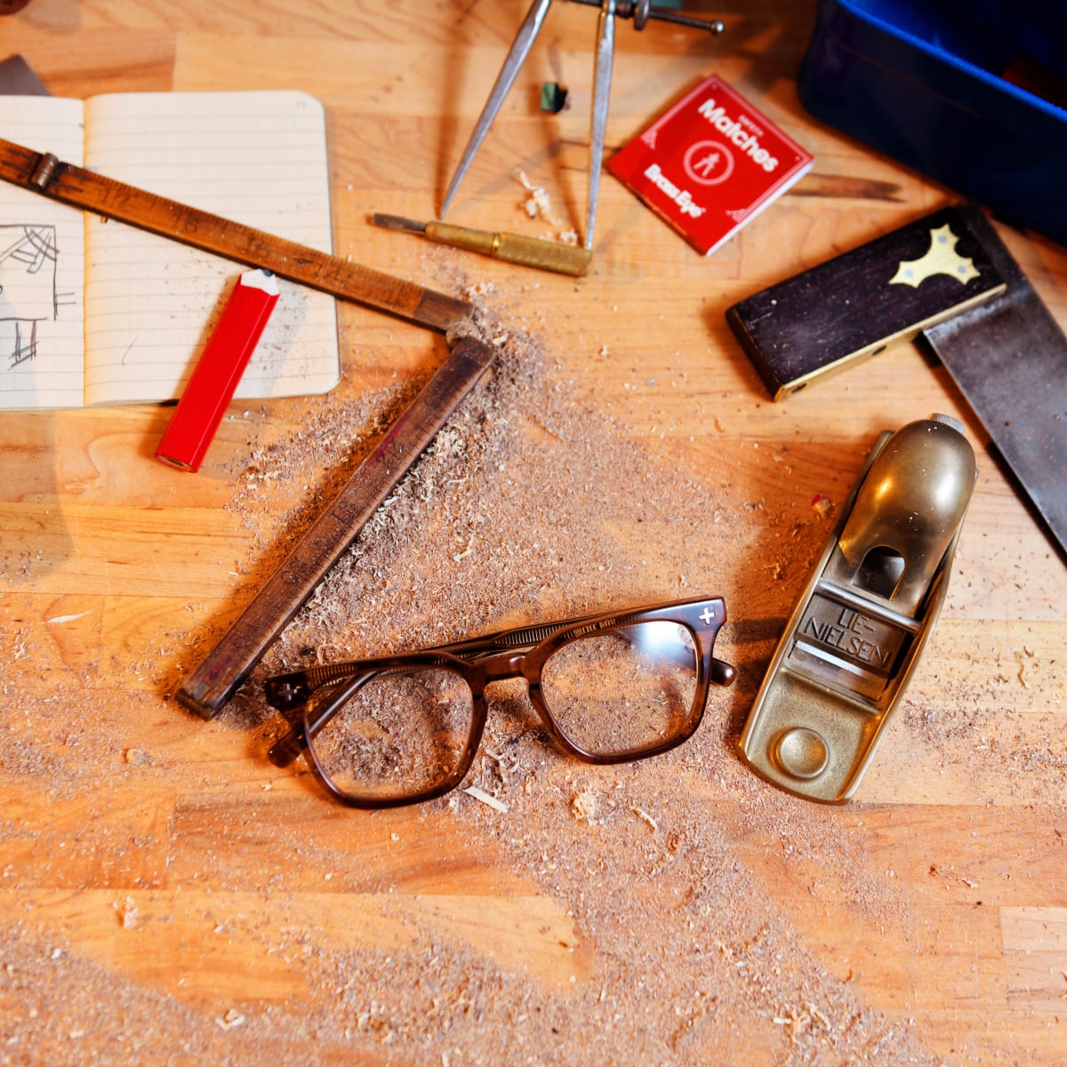 A pair of Tea Brass Eye quality safety glasses rests on a wooden workbench covered with wood shavings, surrounded by woodworking tools including a hand plane, square, compass, and notebook.