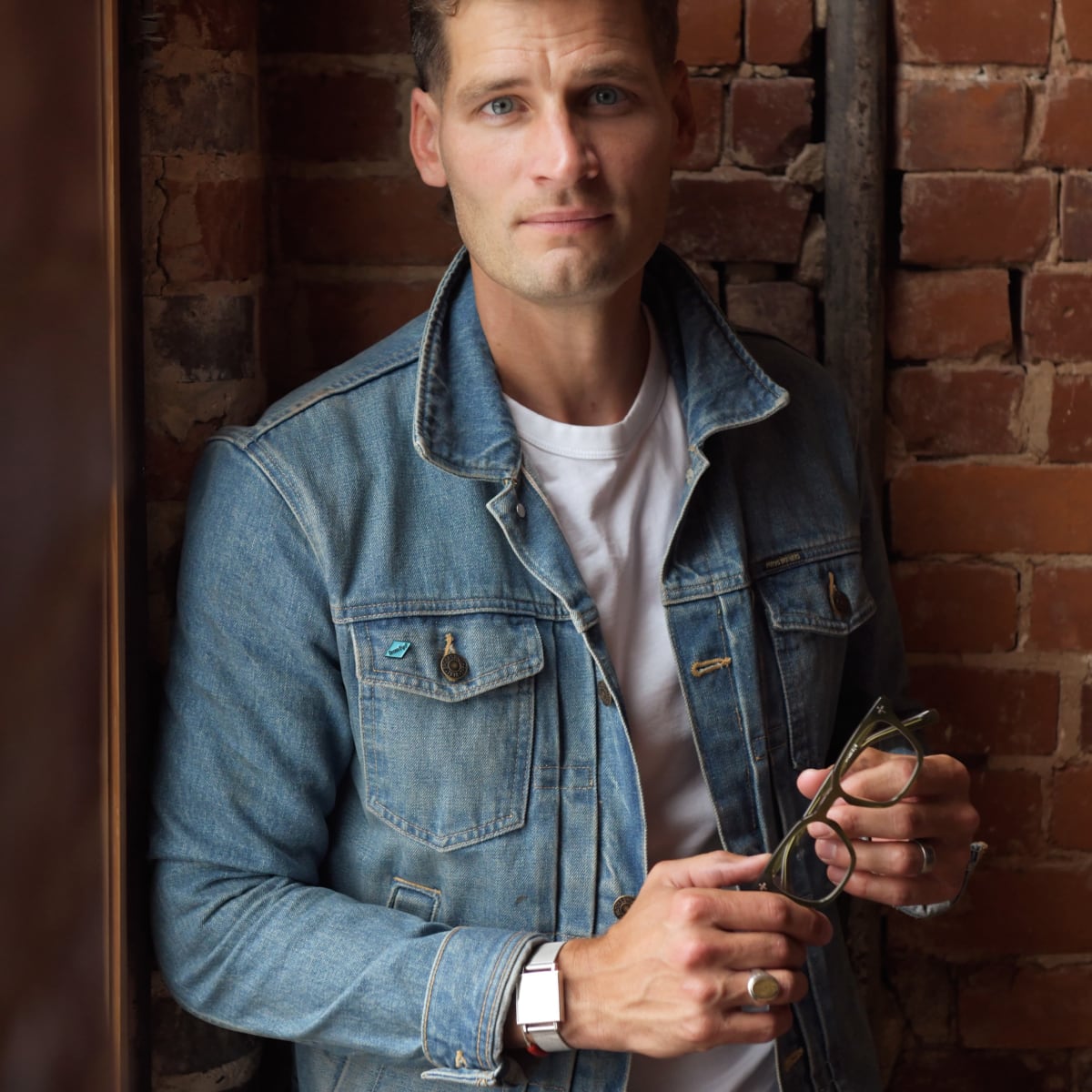 A man wearing a light blue denim jacket over a white t-shirt, holding a pair of Brass Eye quality safety glasses, standing against a rustic red brick wall.